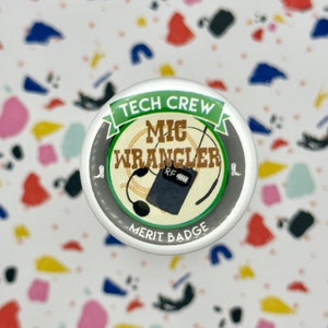 May include: A white button with a green and black design. The button says "Tech Crew Mic Wrangler Merit Badge" with a microphone and a radio transmitter.