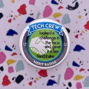 May include: A round, silver button with a blue, green, and white design. The button says "TECH CREW" in a blue banner at the top. The text below says "looked a challenge in the face and gave it a firm handshake" and "MERIT BADGE" at the bottom.