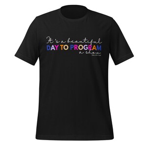 Puede incluir: Camiseta negra con la frase "It's a beautiful day to program a show" en un estilo manuscrito colorido. La camiseta es de cuello redondo y parece estar hecha de un material suave.
