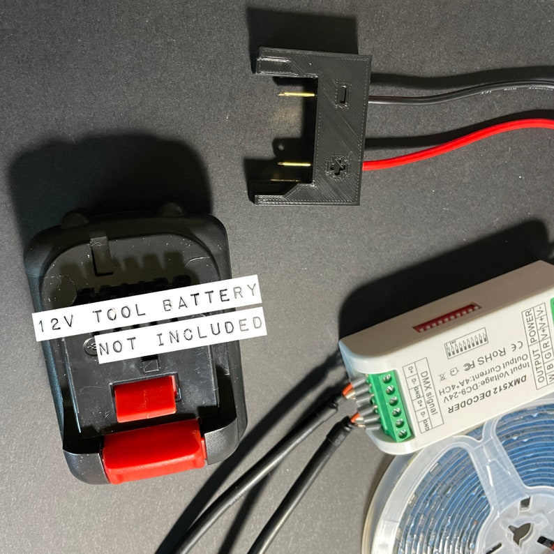 May include: A black battery pack with a red button and a white label that reads "12V TOOL BATTERY NOT INCLUDED". The battery pack is connected to a black and red wire. The wires are connected to a white box with a green terminal block. The box has a label that reads "DMX512 DECODER".