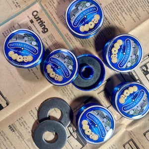 May include: An overhead shot shows several blue and white Pattern Weights tins, labeled "Danish Butter Cookies," alongside metal washers and a sewing pattern. The tins are arranged on a vintage sewing pattern.