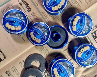 May include: A collection of circular blue metal pattern weights, each featuring a label with the text Pattern Weights and Danish Butter Cookies, alongside two plain grey metal washers, all arranged on a sheet of printed sewing pattern paper.
