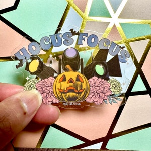 May include: A clear sticker with a whimsical design featuring the words "Hocus Focus" in a retro font. The sticker also includes a jack-o'-lantern, three spotlights, and a floral design. The text "Fly By Night Tees" is at the bottom of the sticker.
