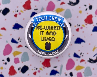 May include: A round, black and white button with a yellow circle in the center. The yellow circle has the text "RE-WIRED IT AND LIVED" in black. The button also has the text "TECH CREW" in blue and "MERIT BADGE" in black.