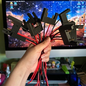 May include: A close-up of a hand holding a bundle of black electrical connectors with red and black wires. The connectors have small metal prongs and are likely used for electrical wiring.