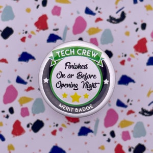 May include: A round, silver button with a green banner that reads "Tech Crew". The button has a black background with white stars and the text "Finished On or Before Opening Night" in white. The bottom of the button reads "Merit Badge" in white.