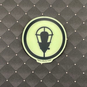 May include: A round, black and white sticker with a glow-in-the-dark background. The sticker features a silhouette of a street lamp with the text "Mighty With All Trades" below.
