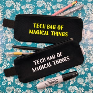 May include: Two black canvas pencil cases with zippers. The top case has the text "TECH BAG OF MAGICAL THINGS" in yellow lettering. The bottom case has the same text in white lettering.