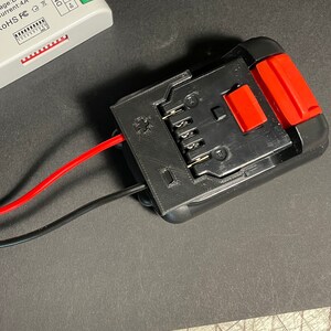 May include: A black battery pack with a red button and six metal contacts. Two wires, one red and one black, are connected to the battery pack.