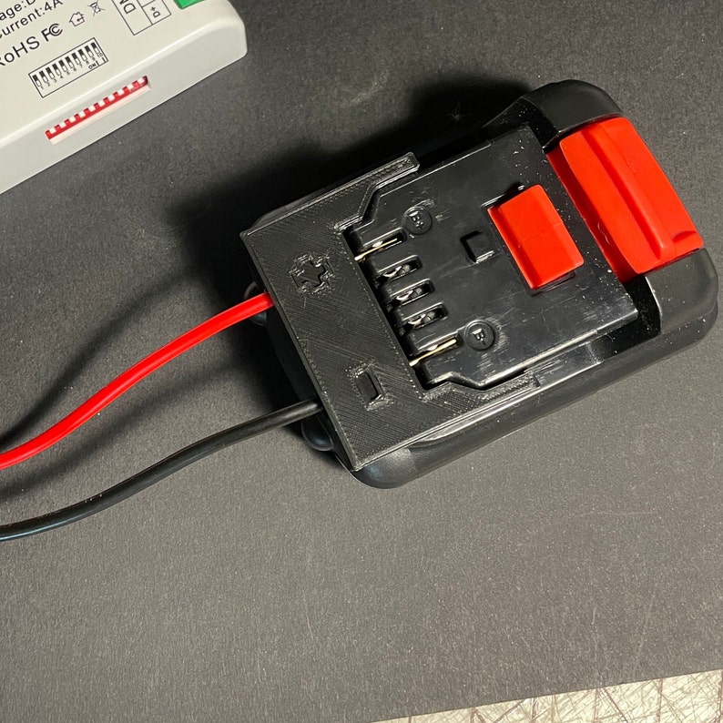 May include: A black battery pack with a red button and six metal contacts. Two wires, one red and one black, are connected to the battery pack.