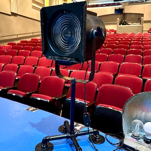 May include: A black vintage theater spotlight on a black pipe stand with a four-footed base. The spotlight is positioned on a blue surface in front of rows of red theater seats. The spotlight has a large lens and a power cord.