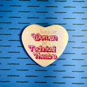 May include: Heart-shaped button with the text "Support Women in Technical Theatre" in pink and iridescent glitter. The button is off-white with glitter and is set against a blue background with black horizontal lines.