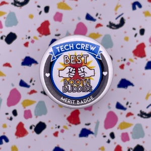 May include: A white button with a black border and blue ribbon reads "Tech Crew Best Booth Buddies Merit Badge". The button features a red sunburst with two fists bumping, and two white hearts.