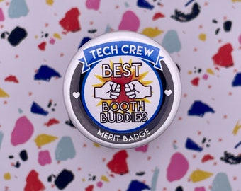 Könnte beinhalten: Ein weißer Knopf mit schwarzem Rand und blauem Band trägt die Aufschrift "Tech Crew Best Booth Buddies Merit Badge". Der Knopf zeigt eine rote Sonnenstrahlen mit zwei aneinanderstoßenden Fäusten und zwei weißen Herzen.