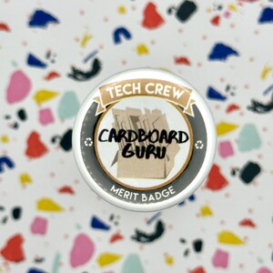 May include: A round merit badge with the text "TECH CREW" and "CARDBOARD GURU". The badge has a black and gold border with a cardboard box graphic in the center. The words "MERIT BADGE" are at the bottom.
