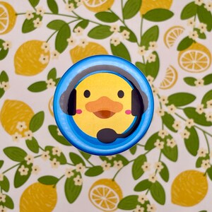 May include: A blue and yellow button with a cartoon duck wearing headphones. The duck has a yellow beak and a light blue background.