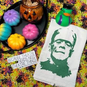 May include: A white kitchen towel with a green silhouette of Frankenstein's face. The text "Because theatre nerds need theatre things" is on a white sticker in the bottom left corner. The sticker also includes the social media handles for "mightywithalltrades.com".