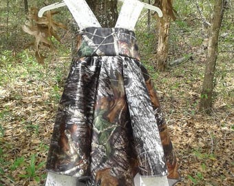 May include: A sleeveless dress featuring a camouflage pattern of brown, green, and grey forest elements. The bodice and skirt are accented with white lace trim along the straps and the hemline, which has a pointed, layered handkerchief style.
