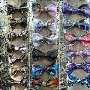May include: A collection of bow ties in various camouflage patterns, including pink, blue, and green. The bow ties are displayed on a burlap background.