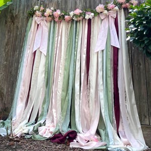 May include: A decorative backdrop with flowing ribbons in pink, green, white, and burgundy. The ribbons are decorated with pink bows and floral arrangements, creating a romantic aesthetic. The backdrop is set against a wooden fence.