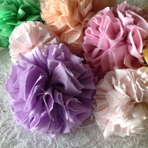 May include: A collection of six fabric flower pom-poms in various pastel colors, including green, pink, peach, lavender, and yellow. The pom-poms are made from soft, ruffled fabric and are arranged in a cluster on a white surface.
