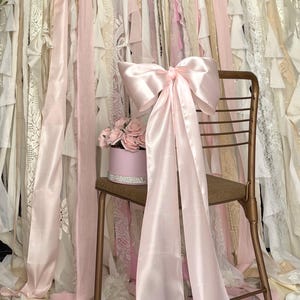 Peut inclure: Un nœud en satin rose clair orne une chaise dorée vintage, avec de longs rubans fluides. Une boîte ronde rose avec une bande argentée contient des roses roses. L'arrière-plan présente des bandes de tissu superposées en rose, crème et blanc.