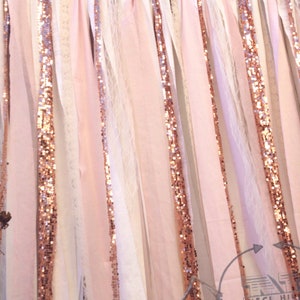 May include: A backdrop made of pink, white, and beige fabric strips with rose gold sequins. The fabric strips are tied together at the top and hang down in a curtain-like fashion.
