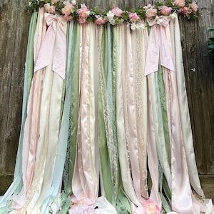 May include: A decorative ribbon backdrop featuring long, vertical strips of satin and lace fabric in shades of sage green, pale pink, and cream. The top is adorned with a garland of artificial pink and white roses and two large fabric bows.