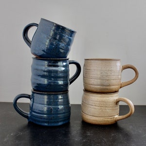 Puede incluir: Cuatro tazas de cerámica hechas a mano. Dos pilas de tazas, una en un esmalte azul oscuro y la otra en un esmalte beige. Cada taza tiene un cuerpo redondeado con un asa. Las tazas azules tienen un diseño texturizado y acanalado.