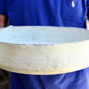 Puede incluir: Un cuenco de cerámica blanco roto hecho a mano con una superficie moteada. El cuenco tiene forma ovalada y un diseño ancho y poco profundo. El borde es ligeramente irregular, lo que añade encanto rústico. El cuenco lo sostiene una persona con una camisa azul.