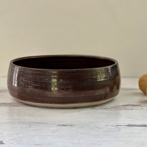 May include: A handcrafted, low-profile ceramic bowl with a rich, dark brown glaze. The bowl has a wide, shallow design with a contrasting white base. Two small, round, light brown objects are to the right of the bowl.