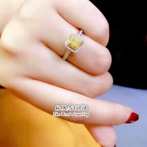 May include: A square-shaped yellow diamond ring with a halo of smaller diamonds set in a silver band. The ring is on a person's finger.