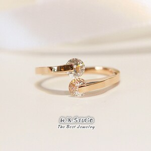 Könnte beinhalten: Ein Ring aus Roségold mit zwei klaren runden Steinen, die in die Band gefasst sind. Der Ring ist so konzipiert, dass er sich um den Finger legt.
