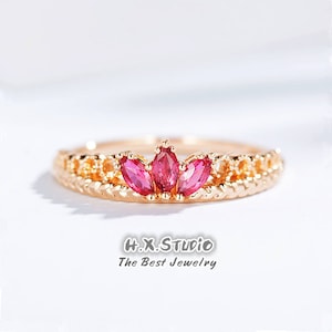 May include: A gold ring with a delicate, braided band and three pink marquise-shaped gemstones set in the centre. The ring is designed to resemble a crown.