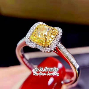 May include: A gold engagement ring with a square-shaped yellow diamond surrounded by a halo of smaller diamonds. The ring is set in white gold and has a delicate band.