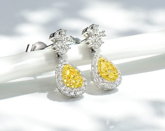 May include: A pair of elegant drop earrings. Each earring features a pear-shaped, bright yellow gemstone surrounded by a halo of smaller, clear stones. The earrings have a silver-colored metal setting and a post-and-stud closure.