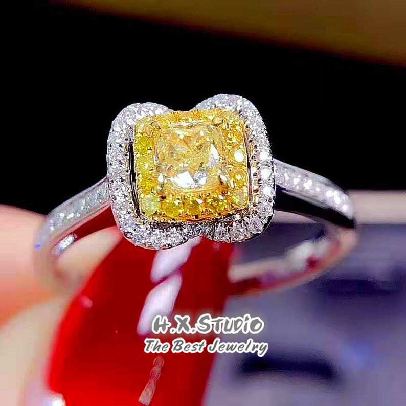 May include: A gold engagement ring with a square-shaped yellow diamond surrounded by smaller diamonds. The ring has a white gold band and a double halo setting.
