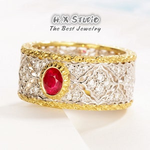 May include: A gold and white ring with a red ruby gemstone in the center. The ring has a delicate, intricate design with small diamonds surrounding the ruby.