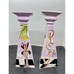 May include: A set of two ceramic candle holders with a pink base and a geometric design. One features a flamingo and the other features a woman in a pink dress.