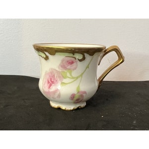 May include: A white teacup with a gold handle and rim, adorned with pink rose floral designs. The cup has a scalloped base and a curved handle. The roses are painted with delicate detail, adding a touch of elegance.