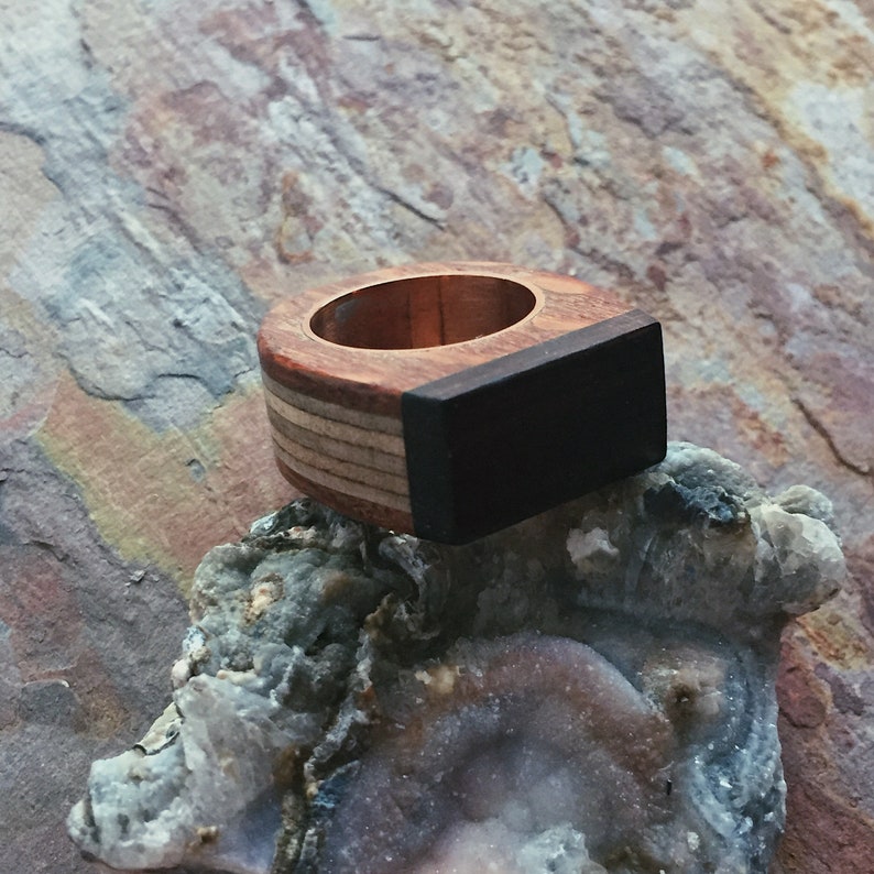 Puede incluir: Un anillo de madera con una incrustaci&oacute;n negra. El anillo est&aacute; hecho de diferentes tipos de madera, incluyendo una madera clara con un patr&oacute;n de veta. El anillo est&aacute; sentado en una roca.
