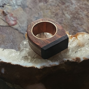 Puede incluir: Un anillo de madera con una incrustaci&oacute;n negra. El anillo est&aacute; hecho de diferentes tipos de madera, incluyendo una madera clara con un patr&oacute;n rayado. El anillo est&aacute; sentado en un cristal blanco.