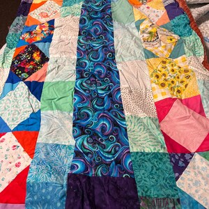 May include: A patchwork quilt featuring a central vertical column of dark blue fabric with swirling purple and teal patterns. The surrounding squares consist of various colourful prints, including sunflowers, floral motifs, geometric shapes, and solid colours.