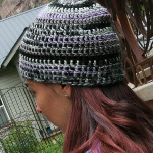 May include: A handmade crochet beanie in shades of black, gray, and lavender. The hat features a textured, open weave design. The beanie is shown on a person with long, auburn hair.