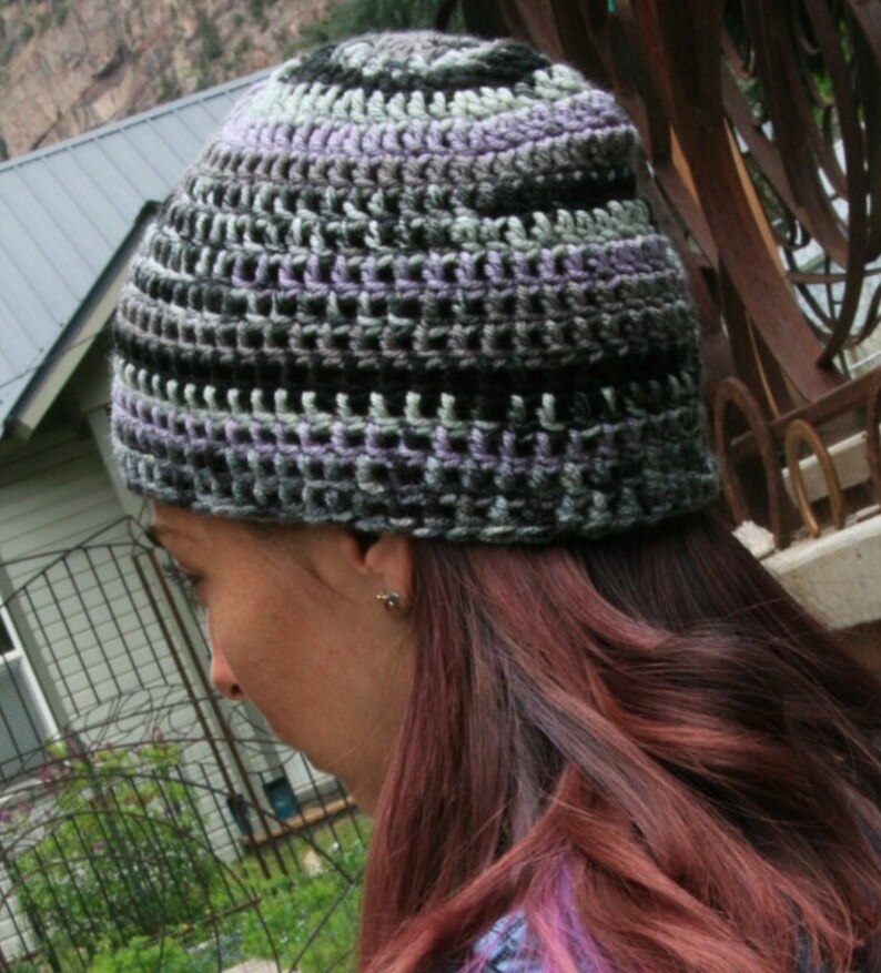 May include: A handmade crochet beanie in shades of black, gray, and lavender. The hat features a textured, open weave design. The beanie is shown on a person with long, auburn hair.