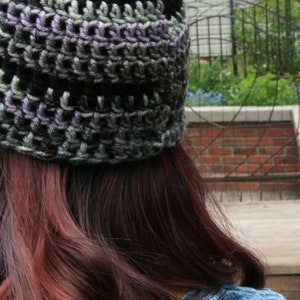 May include: A handmade crochet beanie in shades of gray, black, and purple. The hat is worn, with the back of the head and long auburn hair visible. The beanie's texture is clearly defined, showcasing the intricate crochet work.