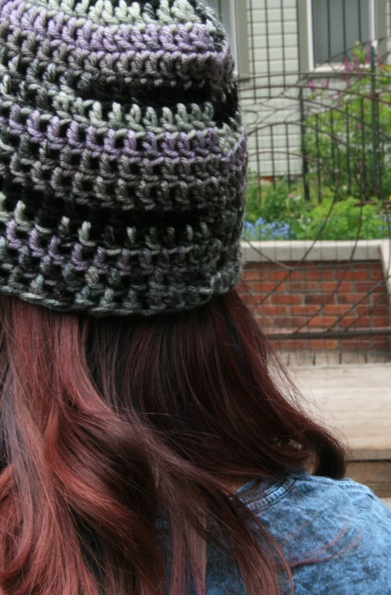 May include: A handmade crochet beanie in shades of gray, black, and purple. The hat is worn, with the back of the head and long auburn hair visible. The beanie's texture is clearly defined, showcasing the intricate crochet work.
