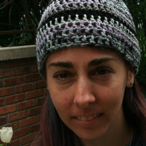 May include: A handmade crochet beanie in shades of gray, black, and lavender. The hat has a textured, open weave design. The person wearing the hat is smiling, with a brick wall and a white tulip in the background.