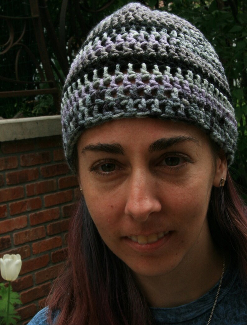 May include: A handmade crochet beanie in shades of gray, black, and lavender. The hat has a textured, open weave design. The person wearing the hat is smiling, with a brick wall and a white tulip in the background.