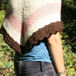 May include: A woman wearing a white, pink, and brown knitted shawl with a blue shirt and dark grey jeans.